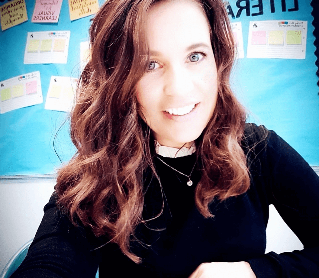A smiling person with long brown hair is in front of a blue wall with sticky notes. They wear a black top and a necklace.